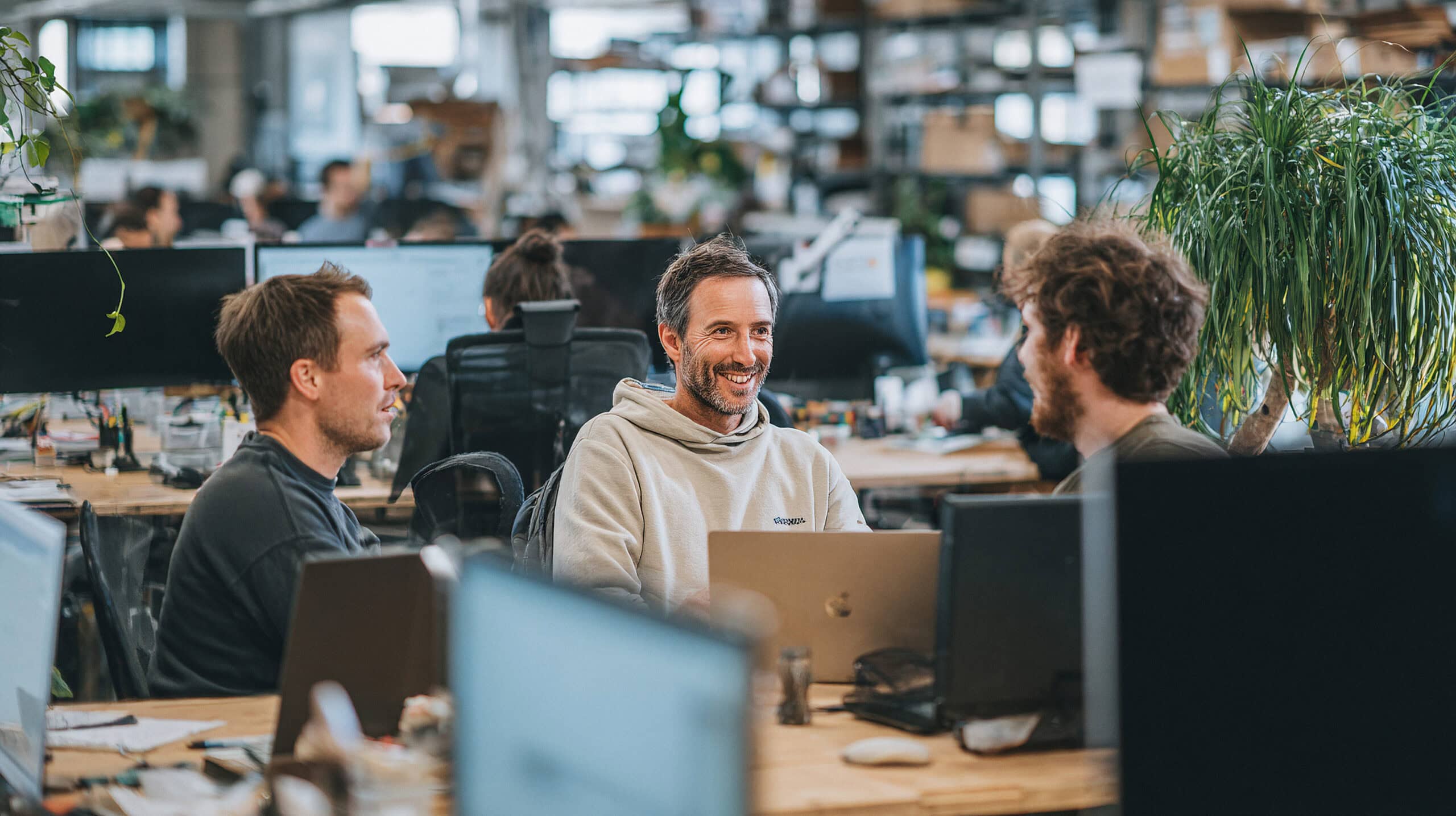Drei Kollegen tauschen sich freundlich am Arbeitsplatz mit Computern aus.